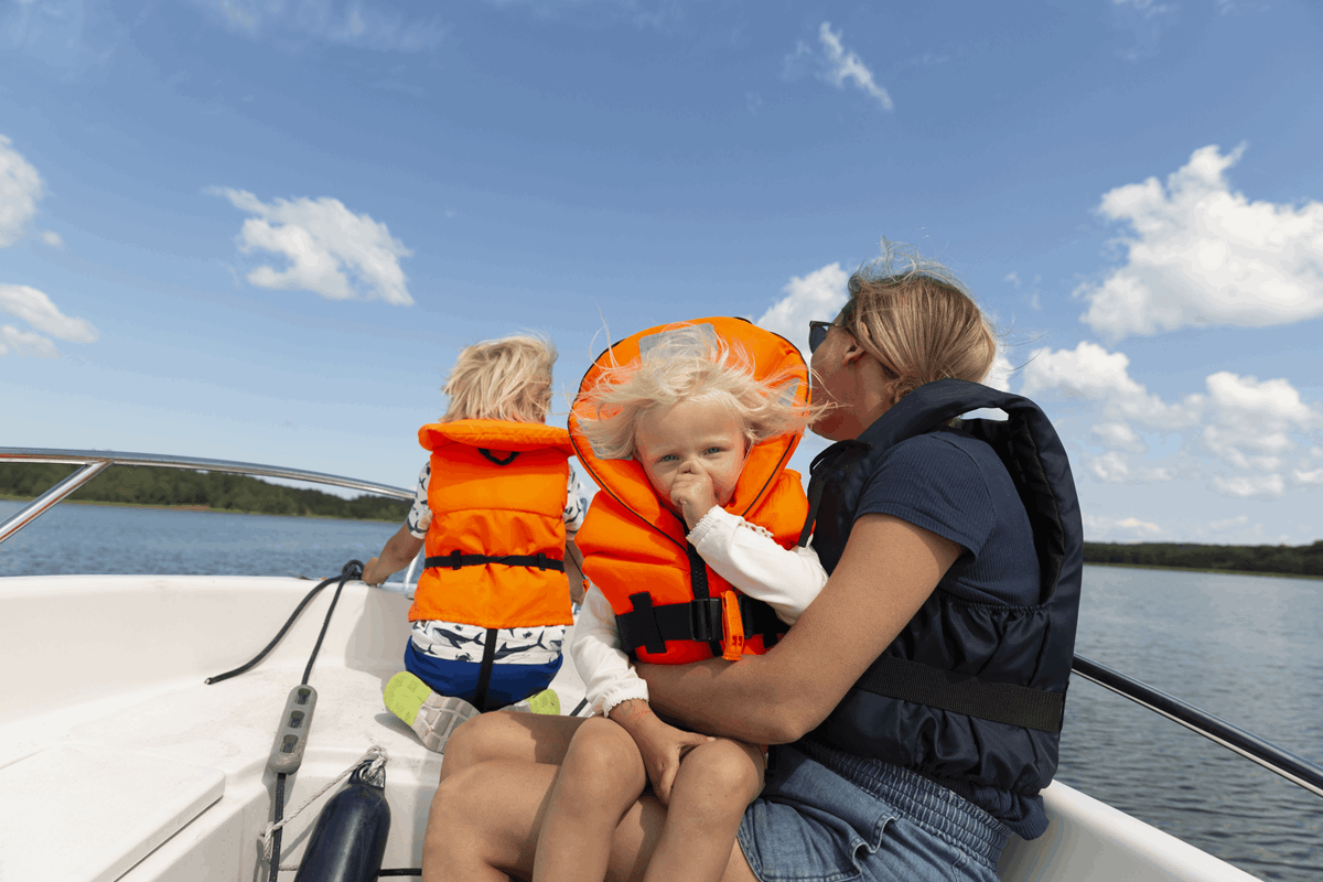Bild på familj som sitter i en båt. En kvinna med två barn och alla har flytväst