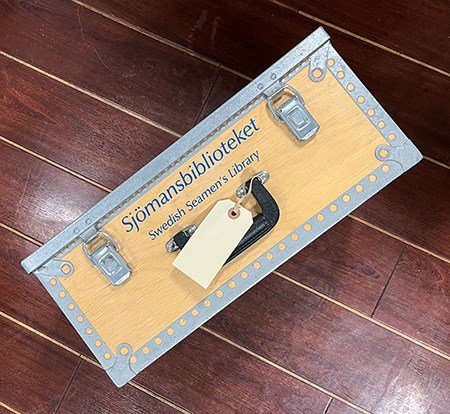 Trälåda med texten Sjömansbiblioteket står på brunt skeppsgolv