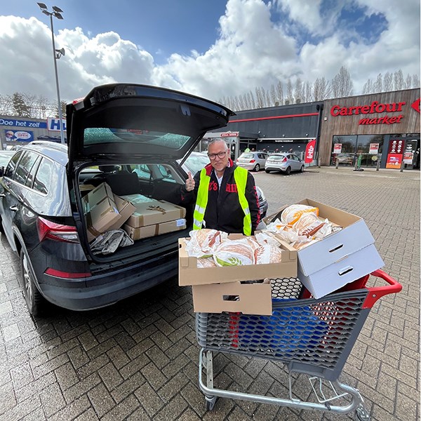 En man står vid en bil med öppen baklucka och en kundvagn med varor. I bakgrunden syns en större butik.