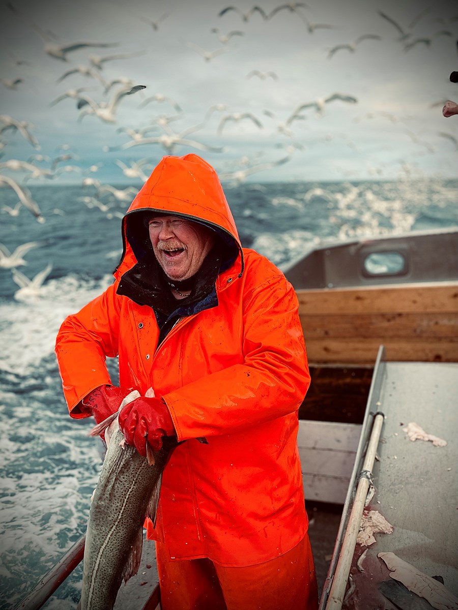 En fiskare i orange regnrock håller i en fisk, i bakgrunden måsar