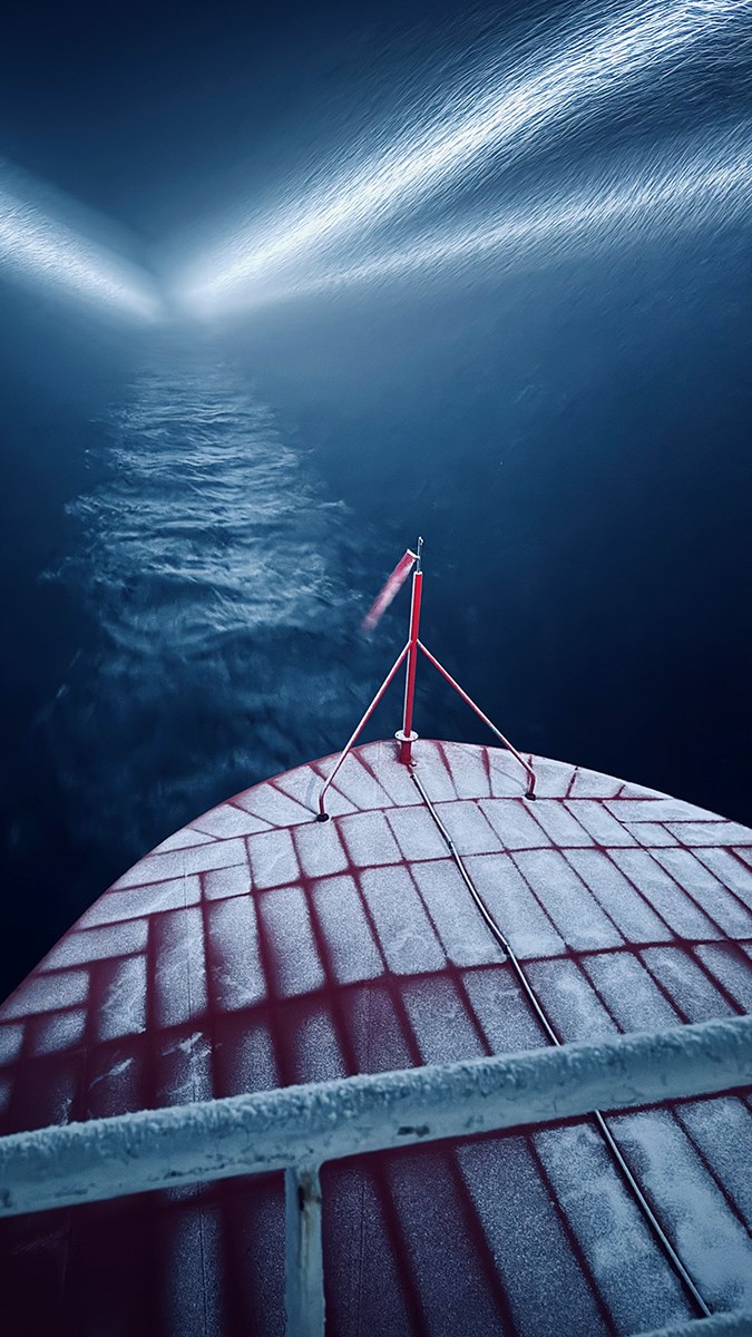 A ship’s bow with a rib‑like pattern of snow on the deck, its headlights illuminating the swirling snow and the path ahead
