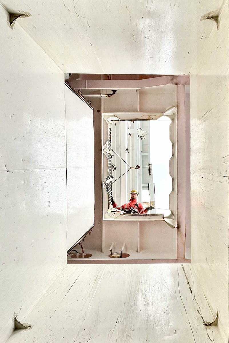 The photo is taken with the camera pointed toward the sky. A worker in a high‑visibility jacket and helmet looks down into a shaft