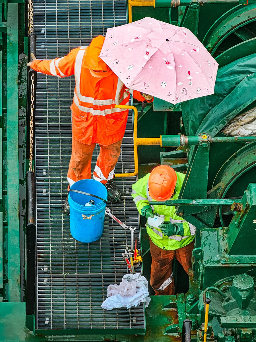 The photo is taken from a slight overhead angle. Two workers in brightly colored high‑visibility clothing are surrounded by tools. One is reading something while the other holds a pink umbrella over him