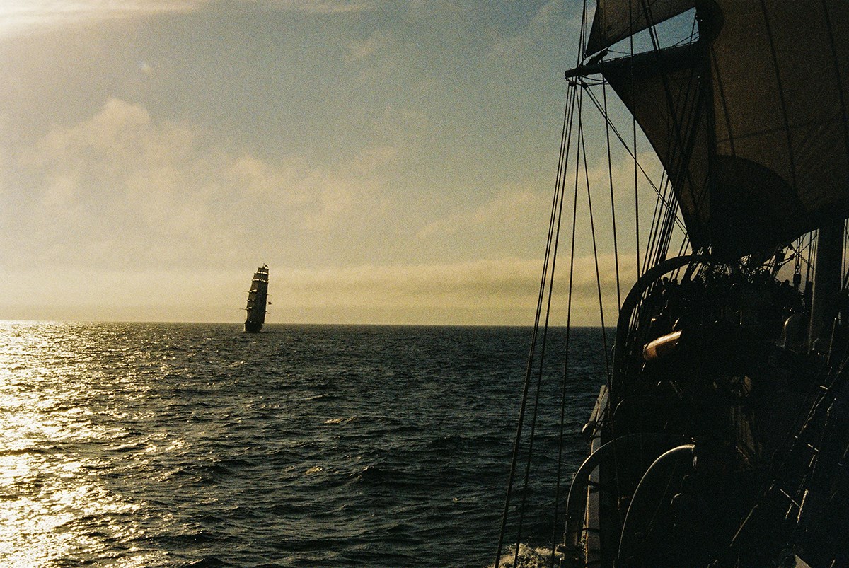On the right, parts of the ship’s own bow and sails are visible. Farther out at sea, it meets another multi‑masted vessel with its sails raised
