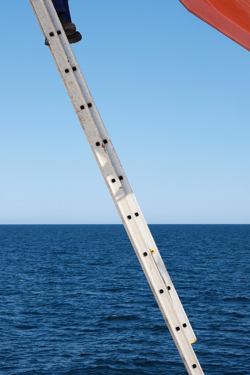 A ladder stretches diagonally across the image. In the upper left corner, a pair of feet can be seen, and in the upper right corner there is an orange‑red object. Near the center of the photo, the sea meets the sky