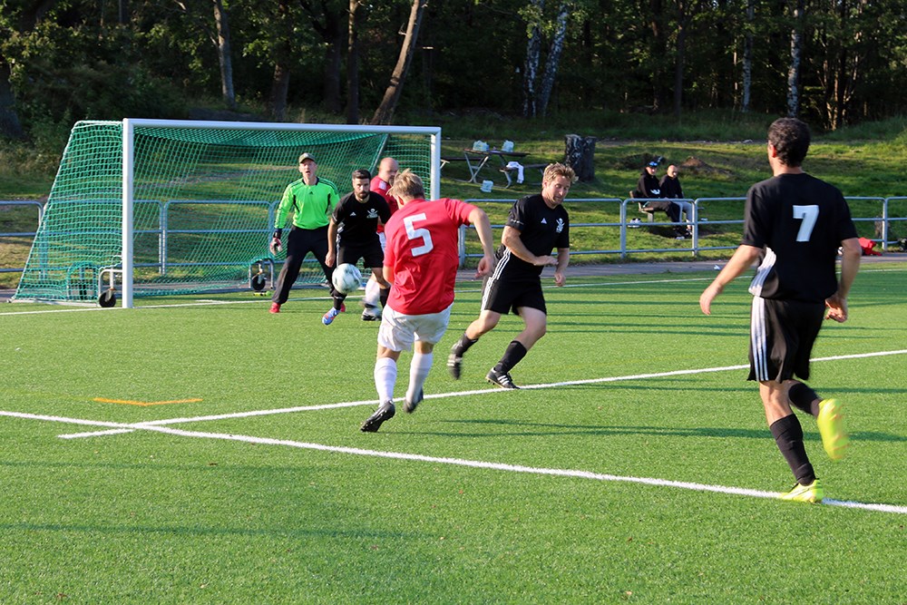 Fotbollsspelare i röd tröja har precis avlossat ett skott mot motståndarens målbur, som täcks av en spelare i svart och målvakten