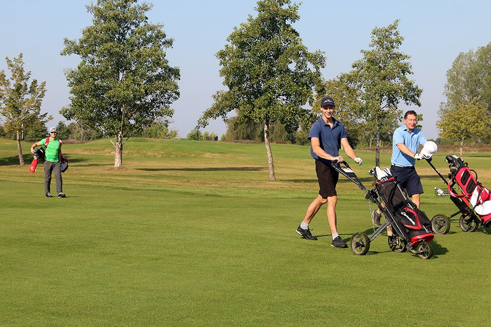 Tre spelare går över golfbanan. De två till höger drar sina golfbagar, medan spelaren till vänster bär sin bag på ryggen