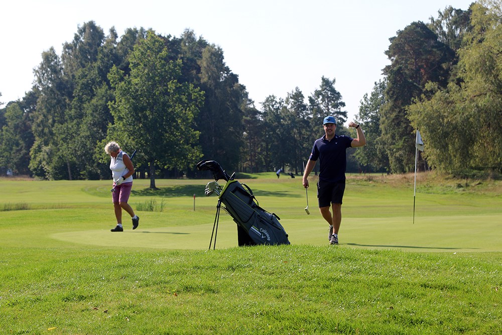 En man går mot kameran och sin golfbag, efter att ha puttat. Spänner armen som i en segergest och ser glad ut. En annan spelare lämnar greenen samtidigt