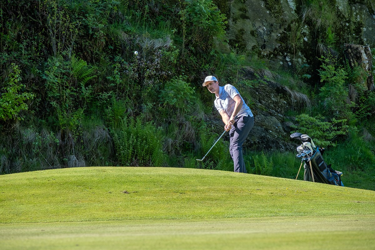 Ensam golfspelare på kulle slår iväg bollen. I bakgrunden ett växtbeklätt berg.